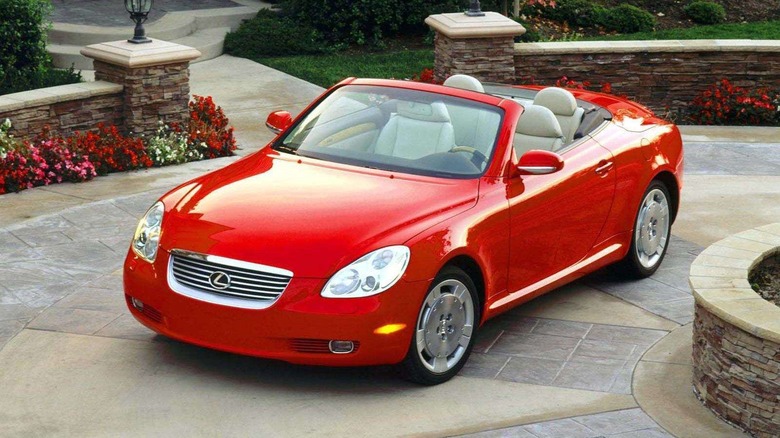 An overhead front three-quarters shot of a bright red 2002 Lexus SC 430 with its top down parked next to a stone wall