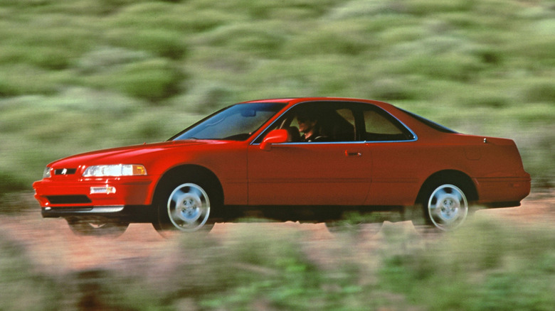 A bright red Acura Legend coupe shot from the front three-quarters angle  driving on a road past blurred greenery