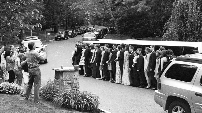 A group of parents take pictures of a group of high school teens dressed up for prom, standing in front of a limousine.