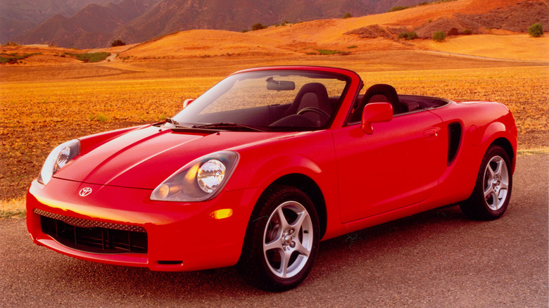 Front three quarters shot of a red 2000 MR2 parked with its top down in front of a yellow field that covers hills