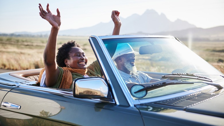 Mature African woman smiling with her hands raised in the air during a scenic road trip with her husband in their convertible