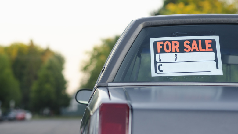 The rear driver's-side corner of an older model GM vehicle, with a For Sale sign taped to the glass, parked on a neighborhood street.