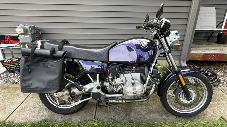 A purple 1992 BMW R100R parked on the sidewalk in front of a grey house.