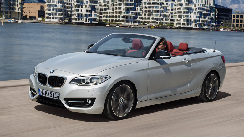 A silver 230i convertible with a red interior driving pn a seaside road in front of white apartments