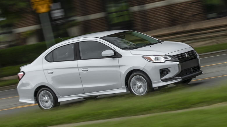 A front three quarters shot of a white mitsubishi Mirage sedan driving  in an urban environment past grass