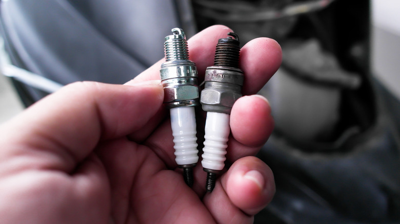 Spark Plug, A hand holds two spark plugs, one new and one used, against a blurred automotive background, highlighting the difference in wear.