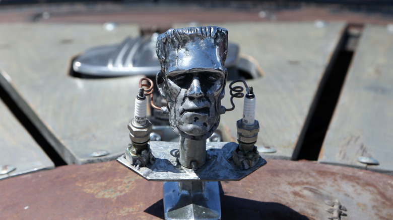 A Frankenstein head with a spark plug on either side, as a hood ornament on a hot rod.