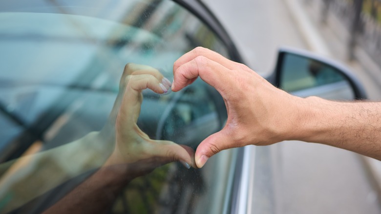A person making a heart-shaped hand gesture, reflected in the car's window.