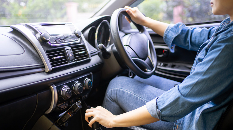 A driver gripping the steering wheel with one hand while using the other to control the manual transmission lever.