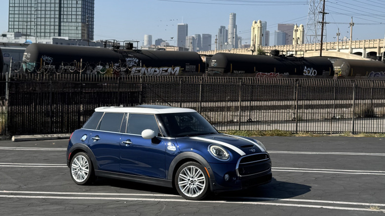 A front three quarters shot of my blue and white 2017 Mini Cooper S four-door hardtop parked in a lot in front of a freight train and a view of the downtown LA skyline