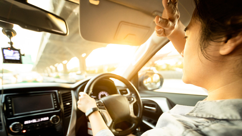 A woman struggling to cover the evening sun blinding her while she tries to drive her vehicle.