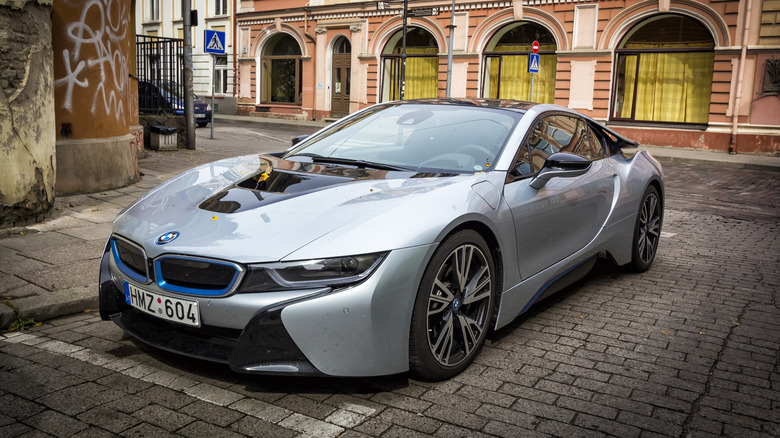 A silver BMW i8 shown from front left parked on a brick lot in front of graffitied pillar and storefront with yellow curtains in Lithuania