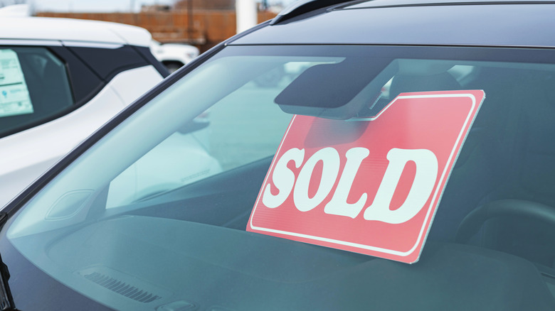 A close-up of a gray car at a dealership with a red sold window sticker.