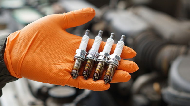 A hand wearing an orange glove holding four used spark plugs.