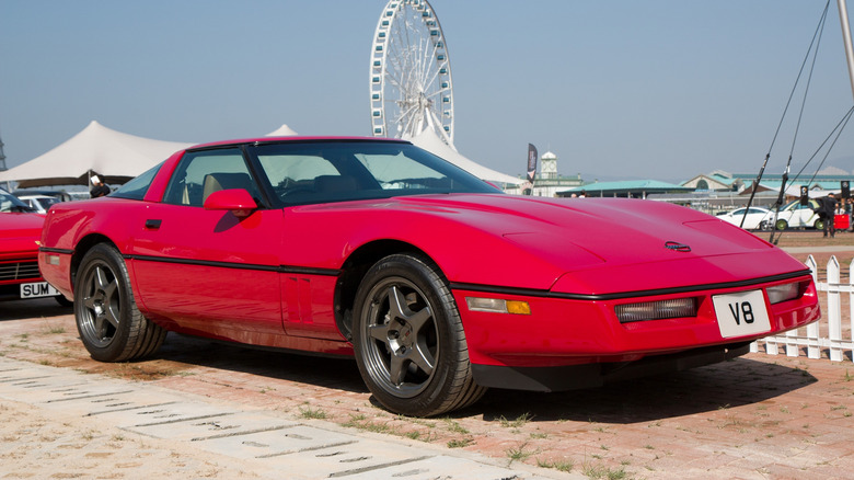 A red C4-generation Chevrolet Corvette parked in front of a white picket fence with a Ferris wheel in the background