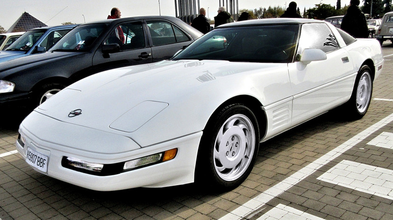 A white C4 Corvette parked in a cobblestone parking lot with numerous other vehicles and people in the background