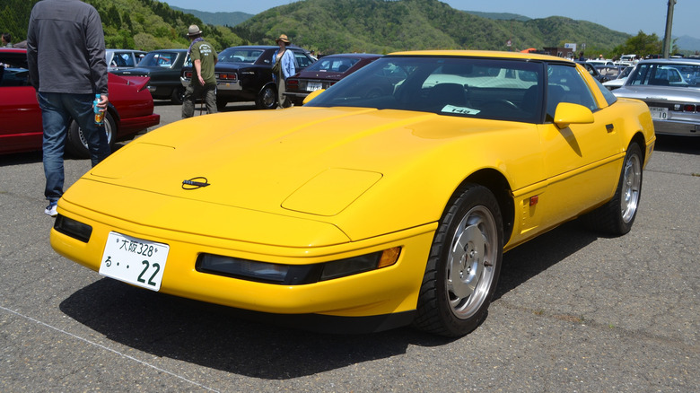 Front-left 3/4 view of a yellow C4 Corvette parked on a paved surface with other cars in the background
