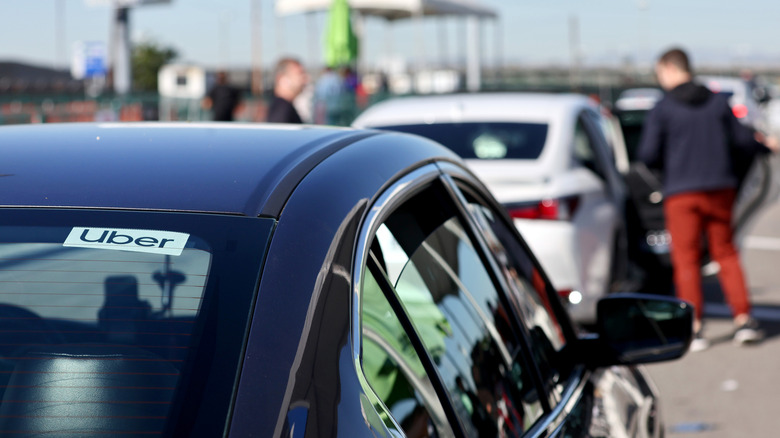 A dark-colored car with an Uber sticker on its rear windshield waits for passengers at an airport pickup area.