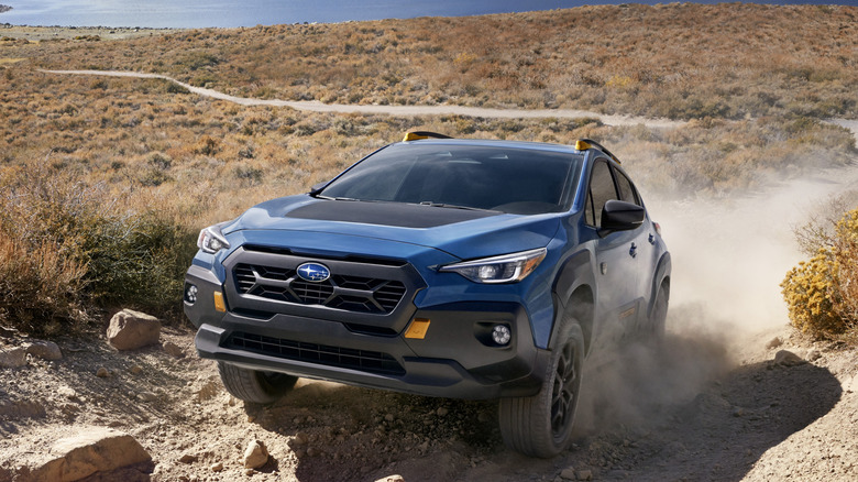 Front three quarters shot of a blue Crosstrek Wilderness climbing a rocky dusty hill in front of a lake and dry brush