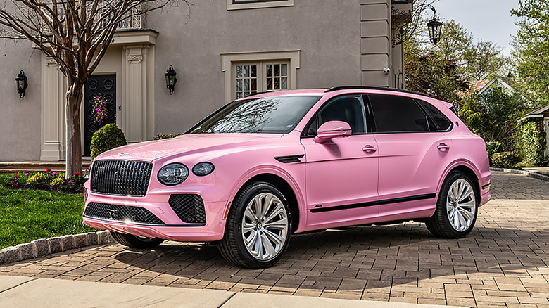 A bright pink Bentayga parked in the driveway of a large gray home with trees