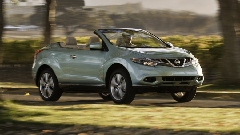 A front three-quarters shot of a minty green Murano CrossCabriolet driving on a rural road past grass and trees