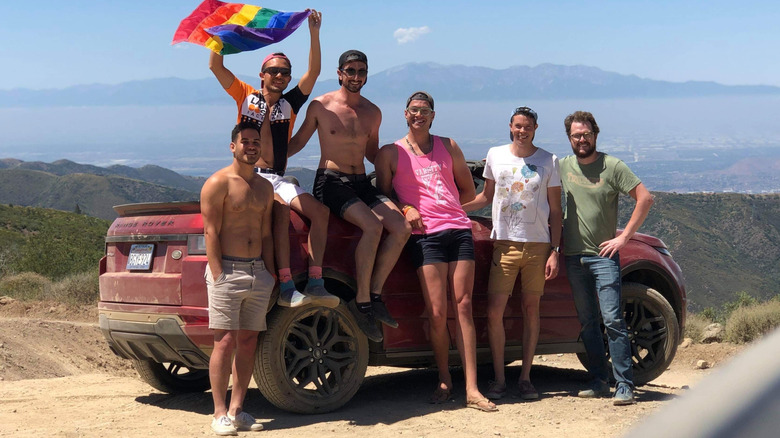 A photo of six queer car enthusiasts in front of a dirty red Range Rover Evoque Cabriolet at the top of a mountain with one waving a Pride flag and a mounatinscape visible in the background