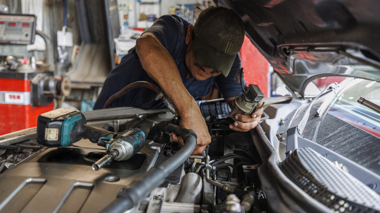 A 57-year-old Latino mechanic leans over an open car hood, meticulously examining and resolving engine components issues using specialized tools.