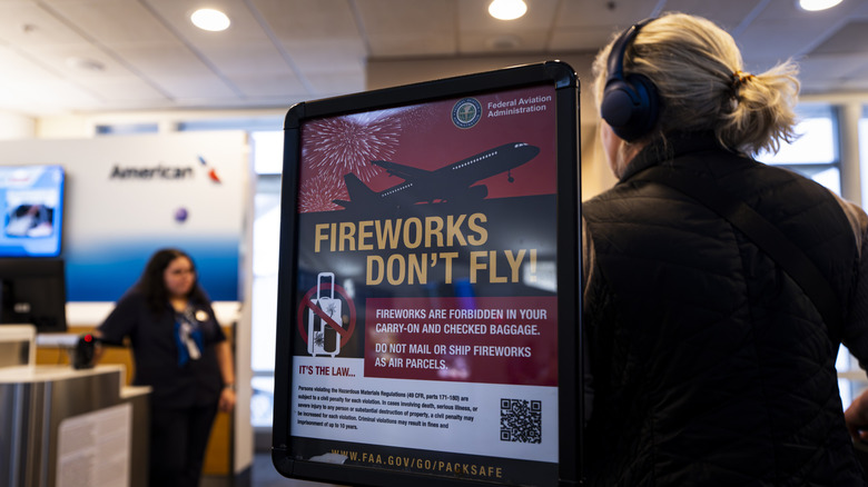 A woman with headphones stands behind a sign that reads "Fireworks Don't Fly" at an American Airlines terminal