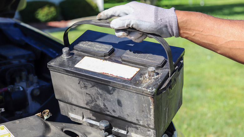 A gloved technician holds up a car battery in front of an open engine bay.