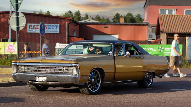 A golden 1970 Imperial LeBaron with black rims on the street with a red building in the background