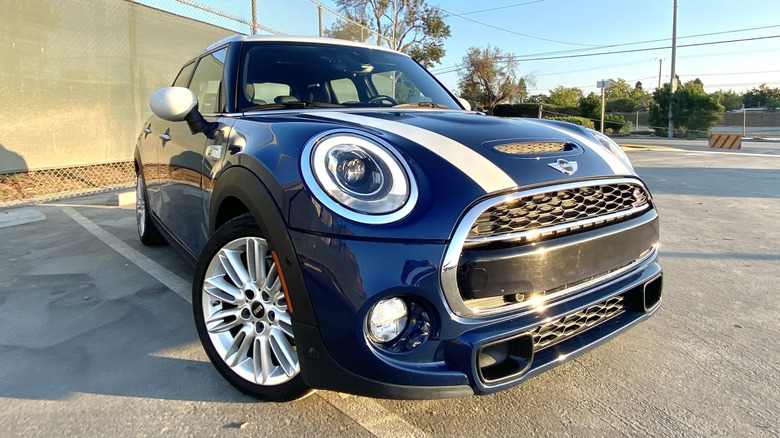 A front three-quarters shot of my blue Mini with white stripes parked in a lot in front of a chainlink fence and a blue sky