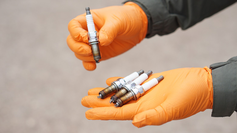 A mechanic in orange gloves holding four used spark plugs