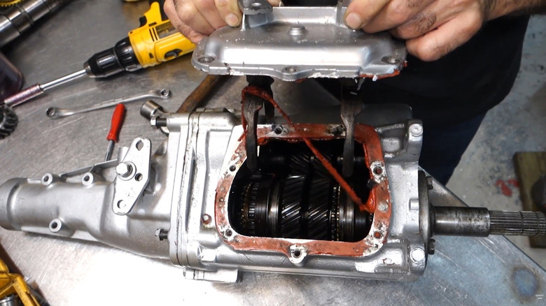 A GM Muncie four speed manual transmission with the side being removed by a man at a metal workbench