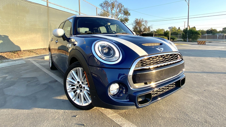 Front three-quarters shot of my blue Mini with white stripes on the hood parked on asphalt in front of a blue sky and a green fence