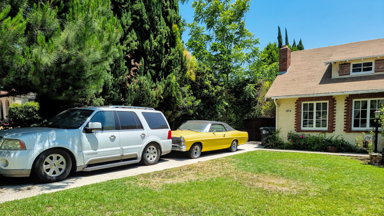 Parked on a gravel driveway next to a traditional house is a large, modern silver SUV and a vibrant, classic yellow car