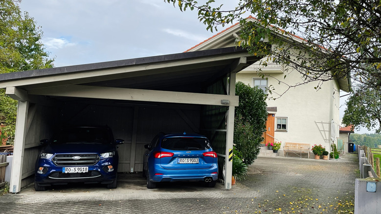 German countryside home with carport, two cars, and a calm yard by a tree-lined driveway.