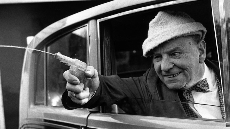a man shoots a water pistol out of his car window with a grin on his face, black and white