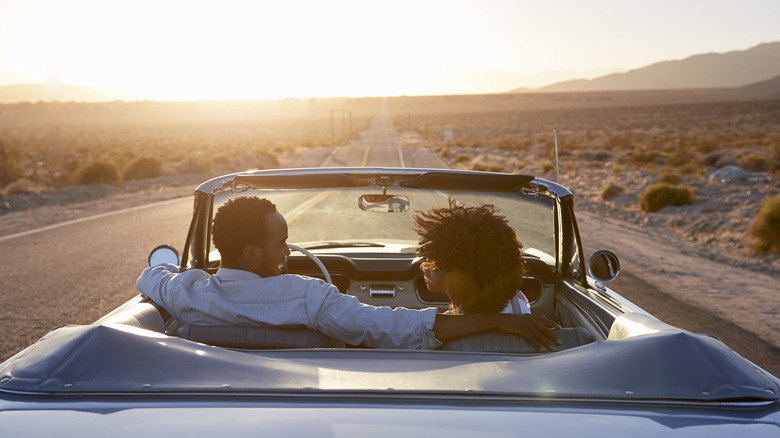 Rear View Of Couple On Road Trip Driving Classic Convertible Car Towards Sunset