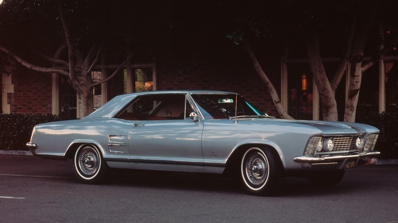 A side view of a light blue 1963 Buick Riviera parked on the street in front of trees
