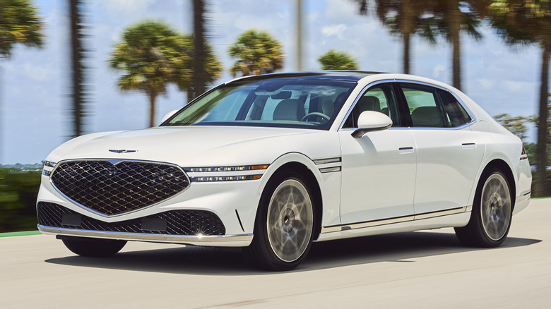 A front three-quarters shot of a white G90 driving past palm trees and a blue sky