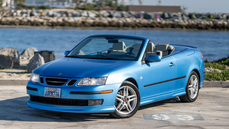 A front three-quarters shot of a light blue Saab 9-3 cabriolet with the top down parked in front of the sea