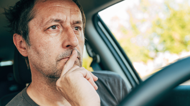 A man with stubble and messy hair concentrating on driving, finger against lips