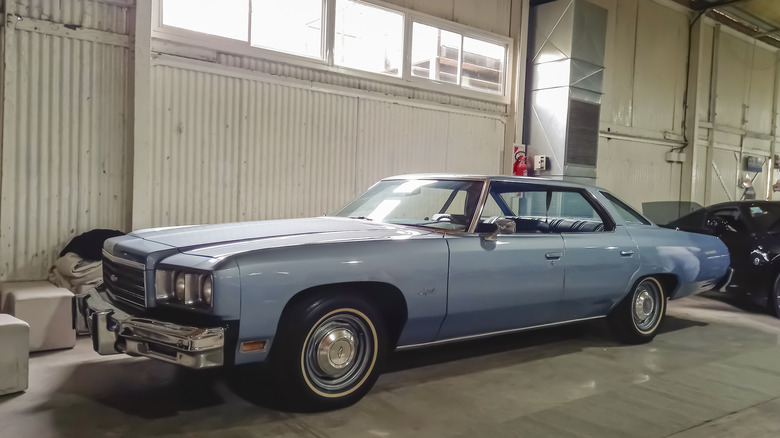 A blue-gray 1970s-era American automobile parked in a garage with other vehicles in the background.