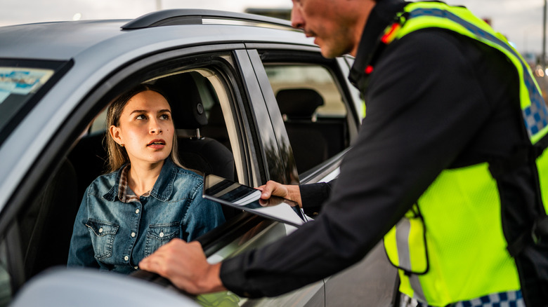 A driver looks through the open window of her car at a police officer who is talking to her with his hand on the window frame.