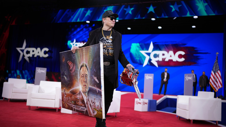 Elon Musk holding a chainsaw and a painting of himself at CPAC