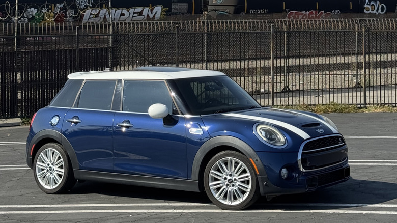 front three-quarters shot of my blue Mini with a white roof and white hood stripes parked in a parking lot in front of a fence and a graffiti'd train