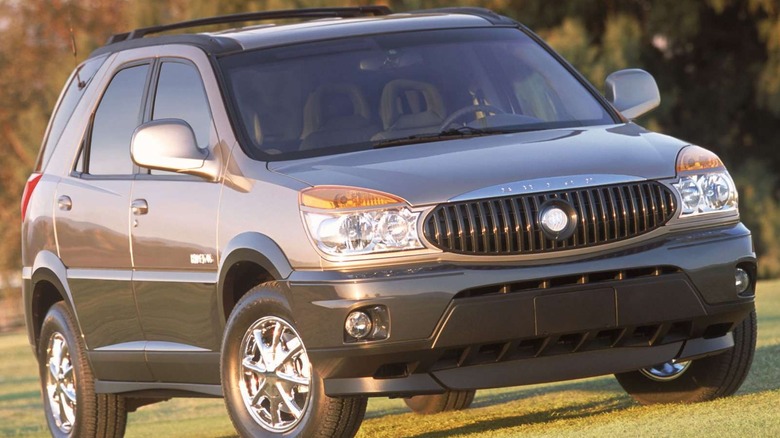 front three-quarters shot of a tan and gray Buick Rendezvous parked on grass in front of blurred trees