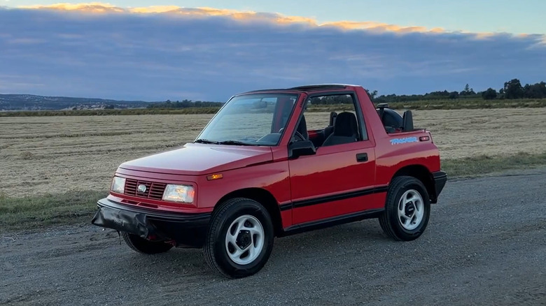 A side shot of a red 1994 Geo Tracker parked in front of a field at dusk