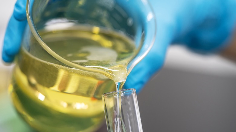 A gloved hand pouring oil from a beaker.