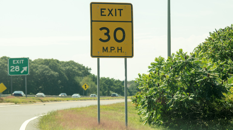 A 30-mph advisory speed sign for highway exit ramp next to a bush with traffic on the road ahead